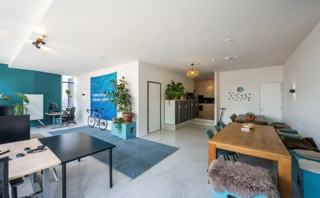 Modern kantoorinterieur aan de James Wattstraat met werkplekken, vergadertafels, planten en een blauwe wand met de tekst "Forecasting Energy Usage".